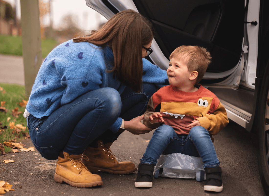 A woman crouches next to a smiling toddler sitting on a portable potty by a car, outdoors on a cloudy day. The woman wears a blue sweater and boots; the child wears a colorful sweater and jeans.