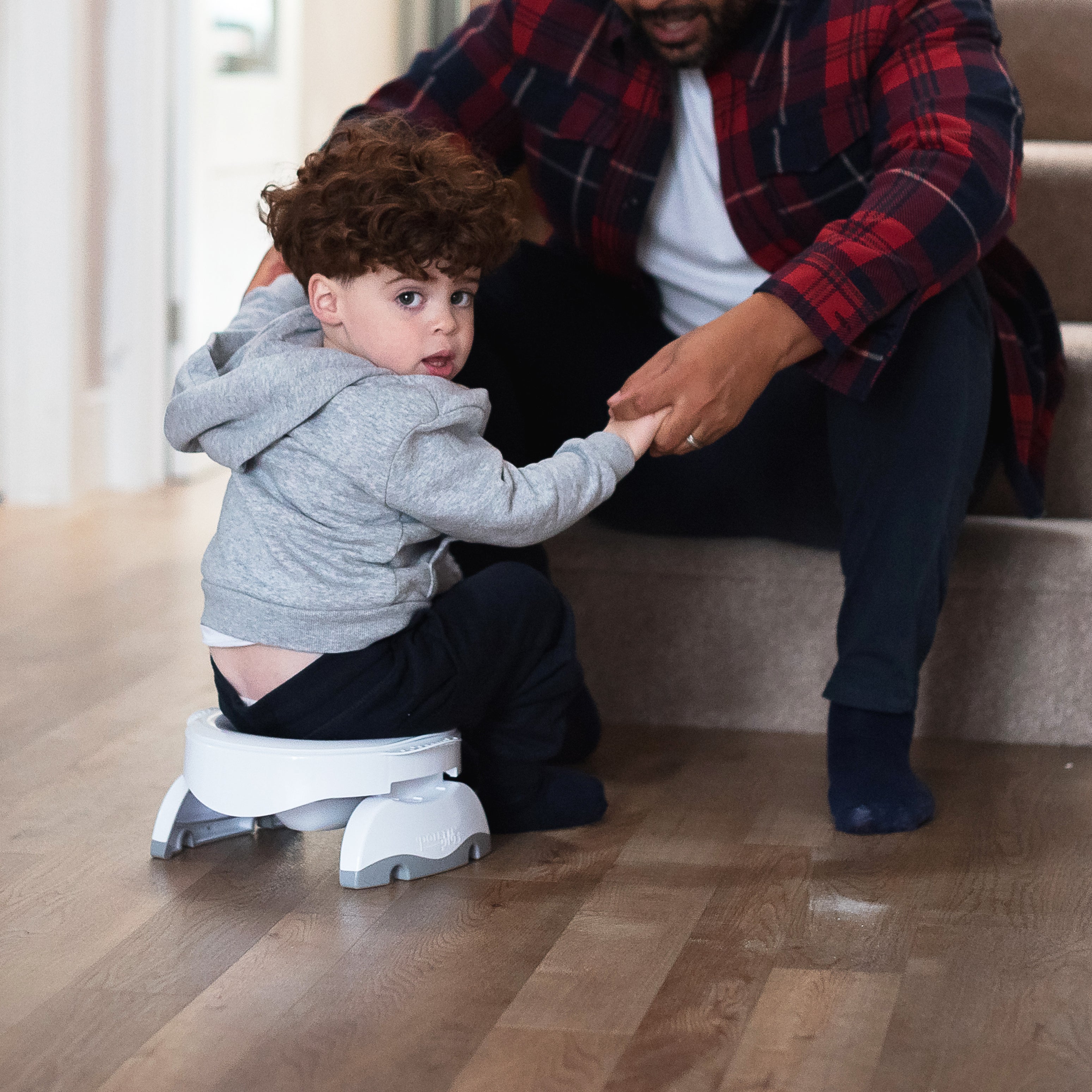 A young child with curly hair sits on a potty training seat on a wooden floor, looking back while an adult in a plaid shirt helps him by holding his hand near a staircase.