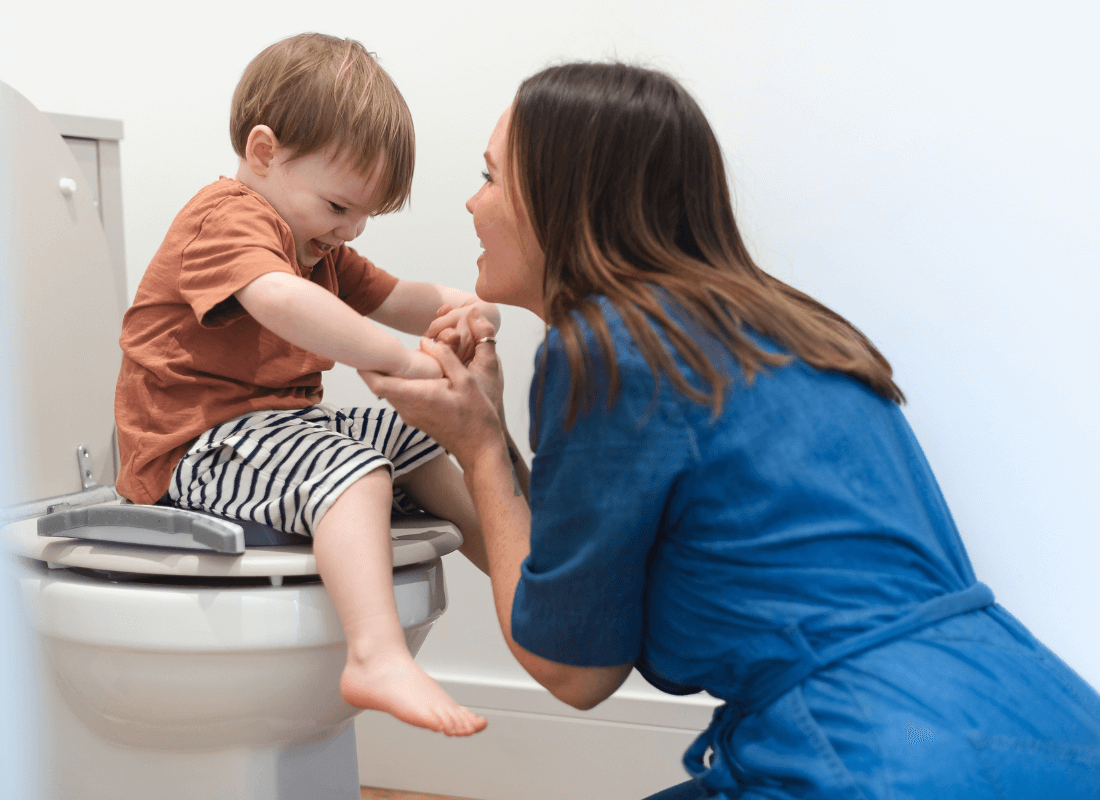 A woman kneels in front of a smiling young boy sitting on a toilet, holding his hands and encouraging him. The boy wears a brown shirt and striped shorts. The setting appears supportive and positive.